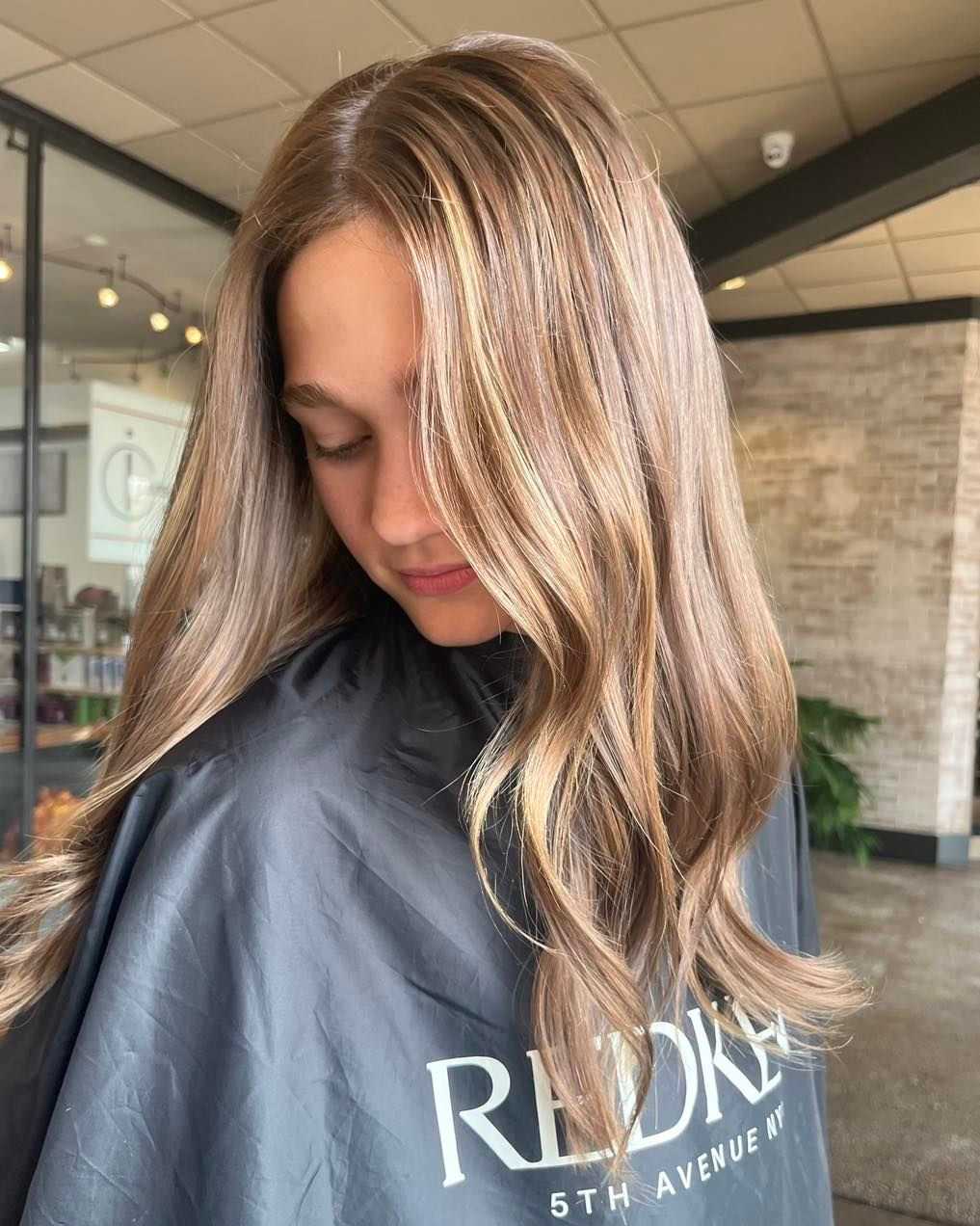 Woman with long, wavy hair and blonde highlights at a hair salon, wearing a black salon cape.